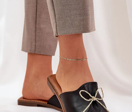 Sandales pour femmes en simili cuir noir avec un nœud décoratif à l'avant, dotées d'un talon plat de 1 cm, idéales pour l'été avec une semelle confortable. Portées avec un pantalon beige et une chaîne de cheville dorée.