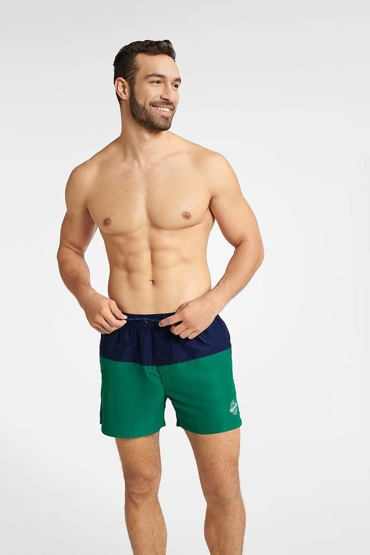 Homme souriant portant un maillot de bain GEM bicolore bleu marine et vert, conçu pour l'été et les activités nautiques, avec une coupe mi-cuisse, une taille élastique et un cordon de serrage. Le maillot arbore un emblème blanc sur le côté gauche avec le mot Summer.