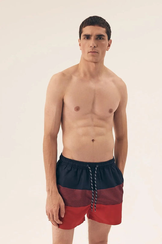 Homme portant un short de bain pour hommes rouge et noir à imprimé tendance, avec une taille élastique et des cordons ajustables. Modèle léger, à séchage rapide, avec une doublure intérieure pour plus de confort. Idéal pour la plage ou la piscine.