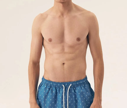 Homme portant un maillot de bain bleu à motifs d'ancres marines, inspiré des plages brésiliennes, alliant style décontracté et élégance tropicale. Parfait pour une journée au bord de l'eau ou une promenade en bord de plage, ce maillot reflète le lifestyle balnéaire moderne et intemporel.