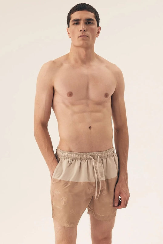 Homme portant un short de bain beige avec des motifs tropicaux subtils, inspiré du style décontracté et festif des plages brésiliennes, idéal pour une promenade en bord de mer ou un moment de détente.