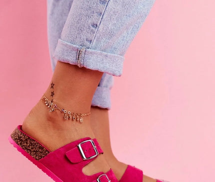 Sandales rose vif avec doubles boucles métalliques, inspirées du style brésilien, portées avec un jean clair retroussé et mises en valeur sur un fond rose tropical. Chaussures confortables idéales pour un look décontracté rappelant l'énergie et le soleil du Brésil.