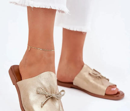 Sandales pour femmes en simili-cuir doré à talon plat, avec un nœud décoratif subtil sur le dessus, idéales pour un look élégant et confortable, portées avec un pantalon blanc à ourlet effiloché.