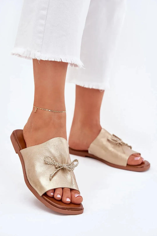 Sandales pour femmes en simili-cuir doré à talon plat, avec un nœud décoratif subtil sur le dessus, idéales pour un look élégant et confortable, portées avec un pantalon blanc à ourlet effiloché.