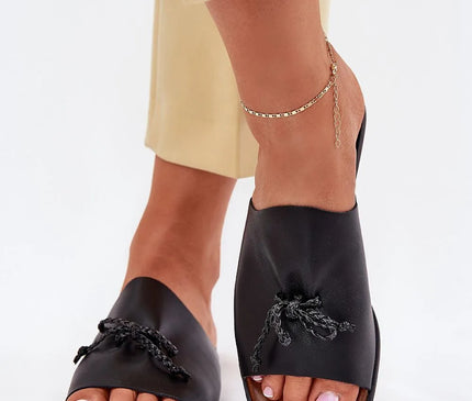 Sandales pour femmes en similicuir noir avec un lacet décoratif subtil, montées sur un talon plat de 1 cm, élégamment portées avec un pantalon beige et un bracelet de cheville doré. Idéales pour un look estival élégant et confortable, adaptées aux robes et pantalons en tissu.