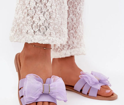 Sandales en cuir écologique pour femmes avec un talon plat de 1 cm, ornées d'un nœud en tulle violet. Idéales pour les tenues estivales, elles s'accordent parfaitement avec des robes, jupes ou pantalons. Présentées avec une tenue blanche en dentelle pour une allure élégante et décontractée.