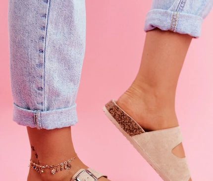 Sandales beige en daim avec semelle en liège, deux larges brides ajustables à boucles argentées portées avec un jean retroussé sur un fond rose, évoquant l'esprit décontracté et ensoleillé du Brésil.