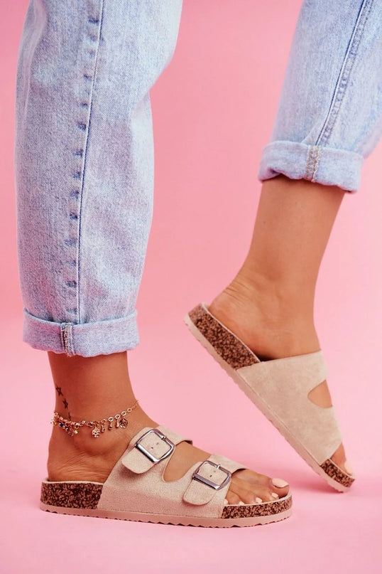 Sandales beige en daim avec semelle en liège, deux larges brides ajustables à boucles argentées portées avec un jean retroussé sur un fond rose, évoquant l'esprit décontracté et ensoleillé du Brésil.