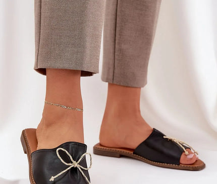 Sandales pour femmes en simili cuir noir avec un nœud décoratif à l'avant, semelle plate de 1 cm, élégantes et confortables, parfaites pour les journées estivales, portées avec un pantalon beige et une chevillière fine.