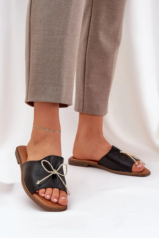 Sandales pour femmes en simili cuir noir avec un nœud décoratif à l'avant, semelle plate de 1 cm, élégantes et confortables, parfaites pour les journées estivales, portées avec un pantalon beige et une chevillière fine.