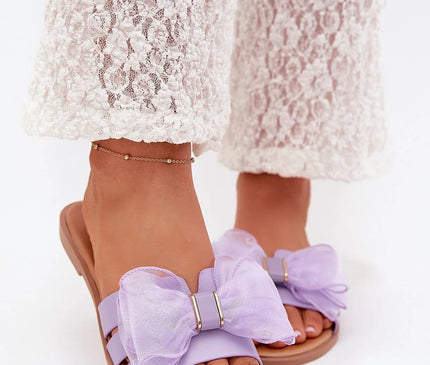 Sandales plates en cuir écologique pour femmes avec un nœud en tulle violet, parfaites pour les tenues estivales, portées avec un pantalon en dentelle blanche.