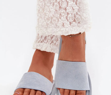 Sandales grises inspirées de la culture brésilienne, élégantes et confortables, sur pieds féminins ornés d'une chaîne de cheville, associées à un pantalon en dentelle blanche, parfaites pour un look décontracté et tropical.