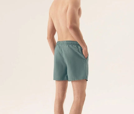 Homme portant un maillot de bain vert au design minimaliste, inspiré de l'esprit détendu et vibrant du Brésil, photographié de dos dans un décor épuré. Idéal pour les journées ensoleillées à la plage ou les activités sportives, ce short incarne l'élégance décontractée des plages de Rio et Bahia.