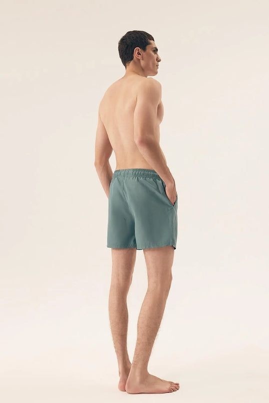 Homme portant un maillot de bain vert au design minimaliste, inspiré de l'esprit détendu et vibrant du Brésil, photographié de dos dans un décor épuré. Idéal pour les journées ensoleillées à la plage ou les activités sportives, ce short incarne l'élégance décontractée des plages de Rio et Bahia.