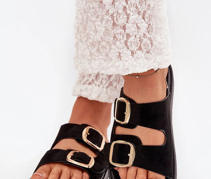 Sandales noires avec larges brides et boucles dorées, inspirées du style décontracté et chaleureux du Brésil, parfaites pour un look chic et confortable, portées avec un pantalon blanc en dentelle pour une élégance naturelle.