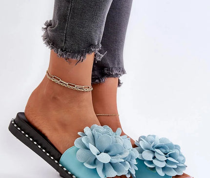 Claquettes pour femmes avec décoration fleurie bleue sur le dessus, semelle noire ornée de petits clous argentés, portées avec un pantalon gris délavé et des chevillères dorées. Idéales pour les journées chaudes et au style tendance.