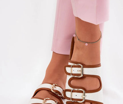 Sandales pour femmes en matériau textile blanc avec des détails en cuir écologique marron, deux sangles ajustables avec boucles dorées, plateforme en liège de 2.5 cm, portées avec un pantalon rose et une chaîne de cheville délicate ; parfait pour un look estival confortable et moderne.