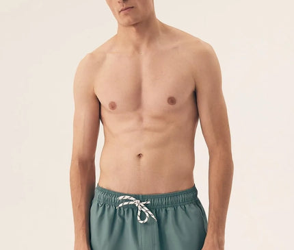 Homme portant un maillot de bain vert de style minimaliste inspiré de l'esprit détendu et vibrant des plages brésiliennes, conçu pour le confort et la liberté de mouvement. Modèle décontracté parfait pour des activités en plein air et des moments de détente au soleil.