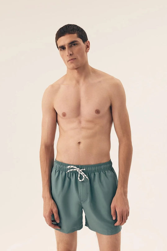 Homme portant un maillot de bain vert de style minimaliste inspiré de l'esprit détendu et vibrant des plages brésiliennes, conçu pour le confort et la liberté de mouvement. Modèle décontracté parfait pour des activités en plein air et des moments de détente au soleil.