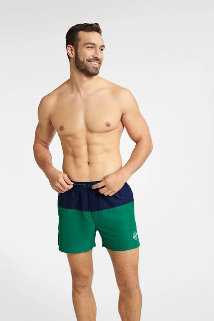Homme souriant portant un maillot de bain GEM bicolore bleu marine et vert, conçu pour l'été et les activités nautiques, avec une coupe mi-cuisse, une taille élastique et un cordon de serrage. Le maillot arbore un emblème blanc sur le côté gauche avec le mot Summer.