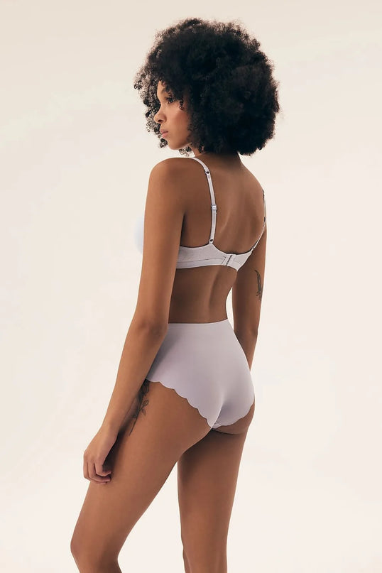 Femme de dos portant une lingerie élégante et épurée s'inspirant de la culture brésilienne, avec des finitions délicates et un design naturel. Taille haute en dentelle ondulée blanche, symbolisant le charme tropical et l'esprit libre des femmes brésiliennes, évoquant les plages de Rio et un style raffiné.