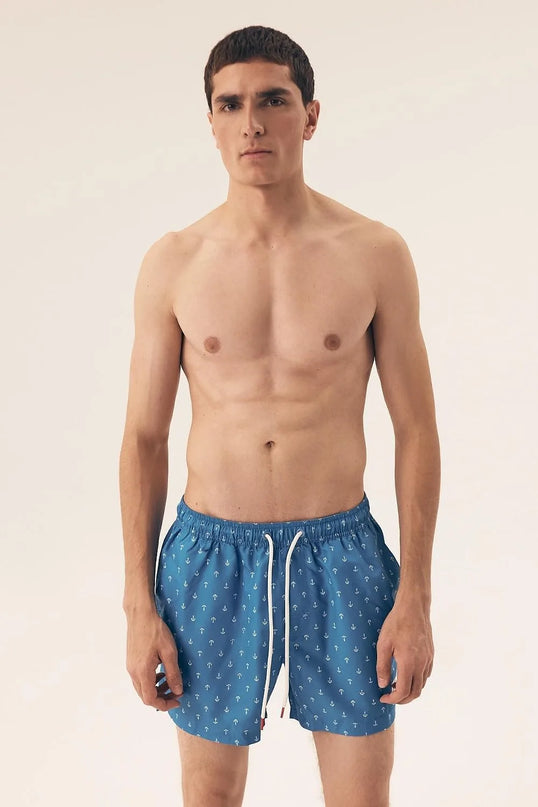 Homme portant un maillot de bain bleu à motifs d'ancres marines, inspiré des plages brésiliennes, alliant style décontracté et élégance tropicale. Parfait pour une journée au bord de l'eau ou une promenade en bord de plage, ce maillot reflète le lifestyle balnéaire moderne et intemporel.
