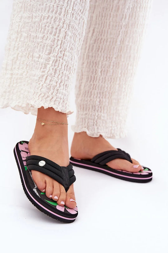 Sandales inspiration brésilienne avec semelle florale noire et rose, brides rembourrées noires pour un confort optimal, portées avec un pantalon blanc en tissu léger, évoquant le style décontracté et les plages animées du Brésil.