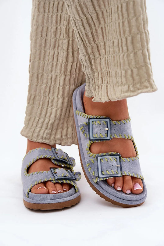 Claquettes pour femmes en suédine écologique grise avec boucles décoratives sur le devant et coutures contrastées jaunes, semelle intérieure profilée pour un confort optimal, présentées portées avec un pantalon beige texturé. Idéales pour les jours d'été et les styles urbains ou de vacances.